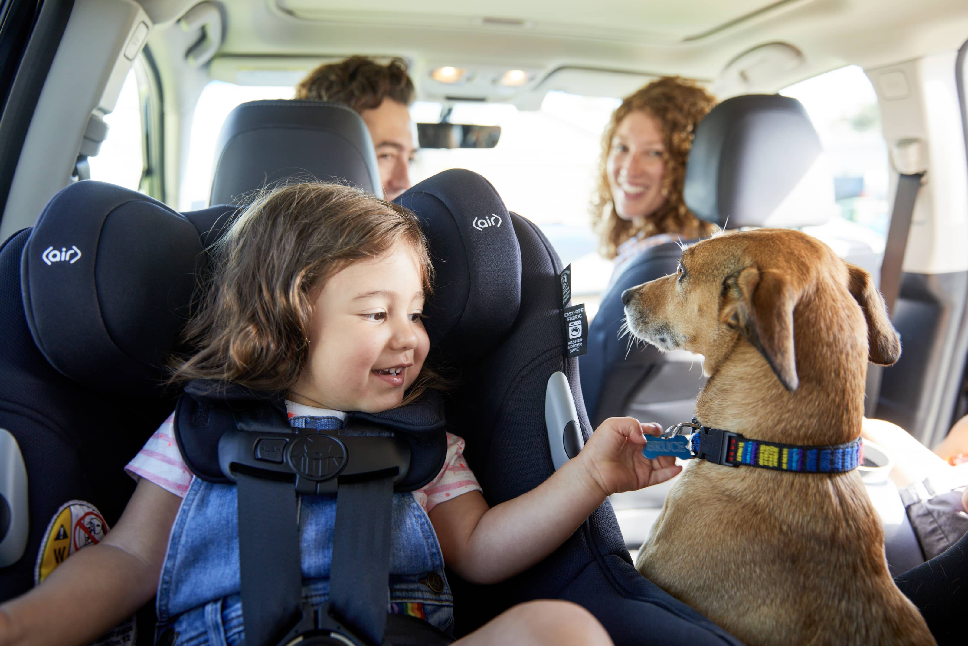 A child sits in a car seat and plays with her dog, Bruce's collar as her parents watch from the front seat.
